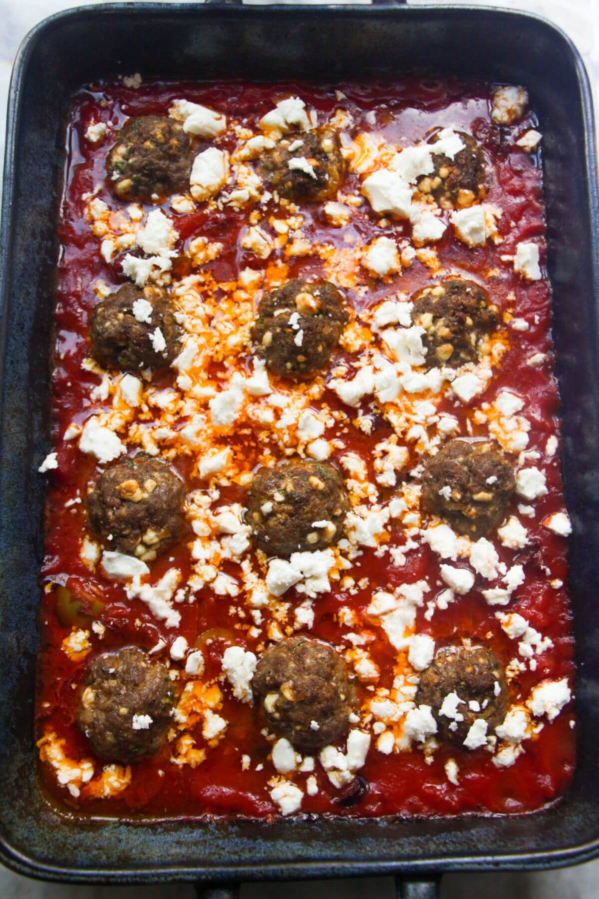 12 meatballs in tomato sauce in a large blue oven dish with feta scattered over the top.