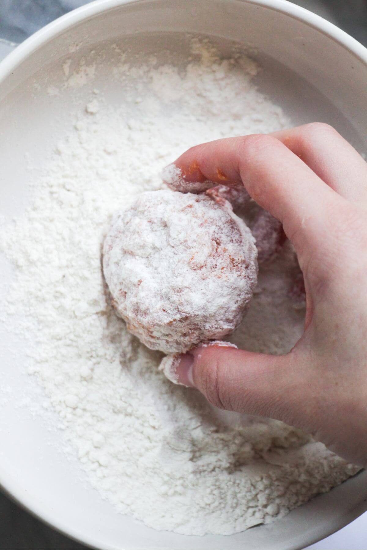 Hand tossing arancini ball in a small bowl of flour.