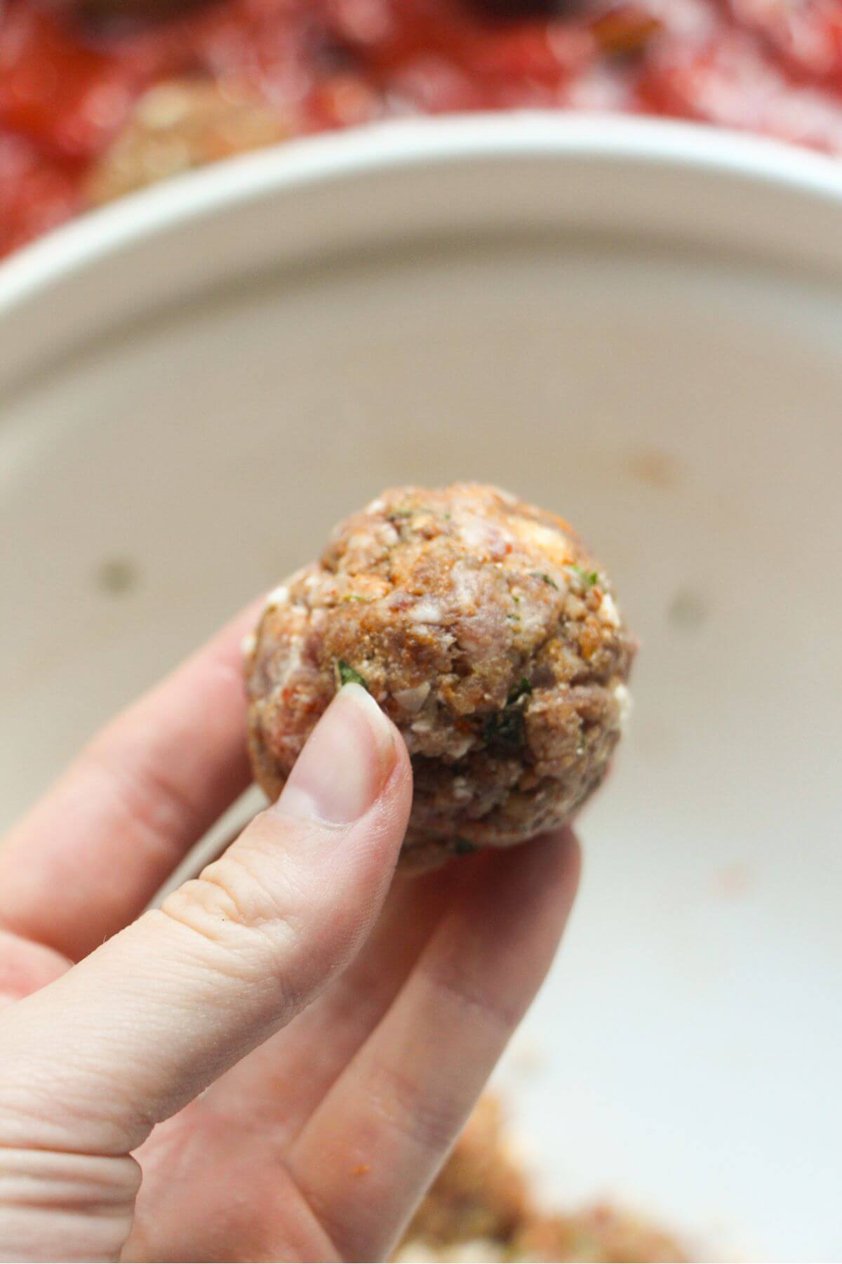 Hand holding a formed meatball, with white mixing bowl in the background.