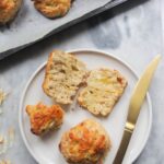 Small white plate with a whole and a sliced open cheese scone, with a gold knife on the side and tray of scones in the background.