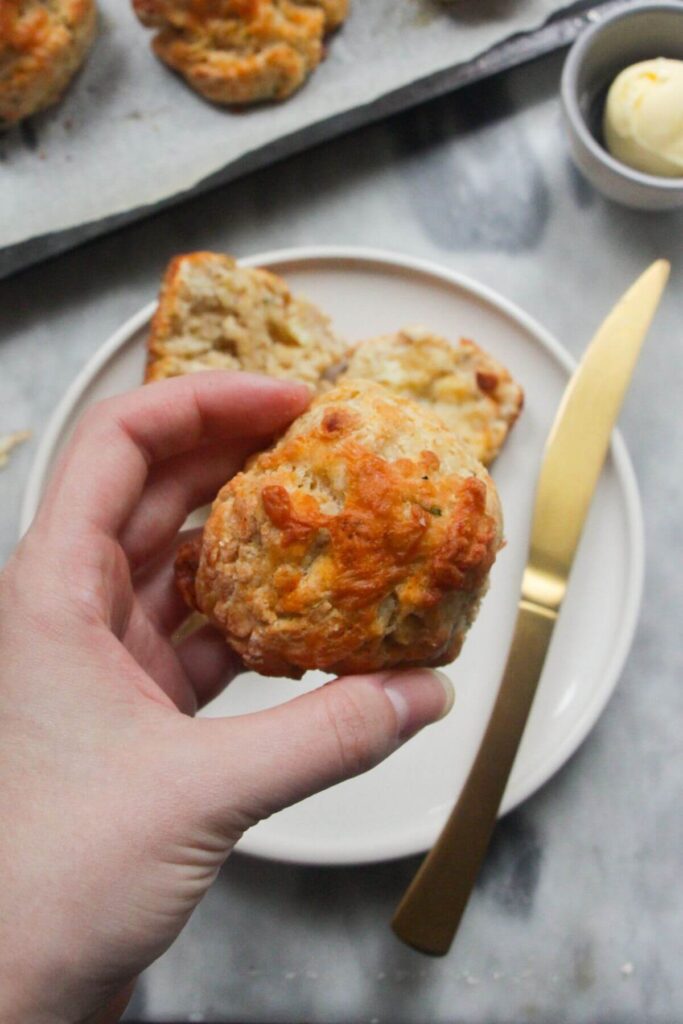 Hand holding up a cheese topped cheese scone, with small white plate with another cheese scone on it in the background.