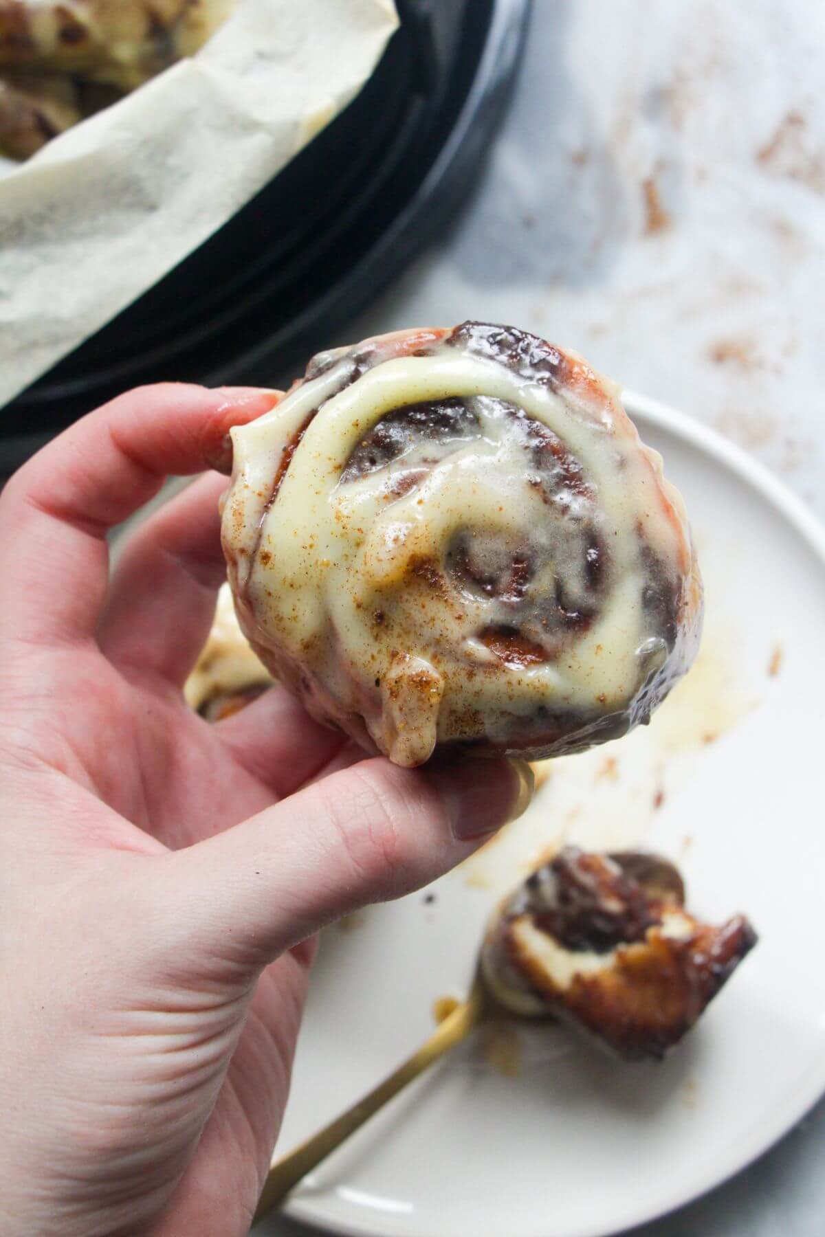 Hand holding up a pumpkin spice cinnamon roll with plate of more rolls in the background.