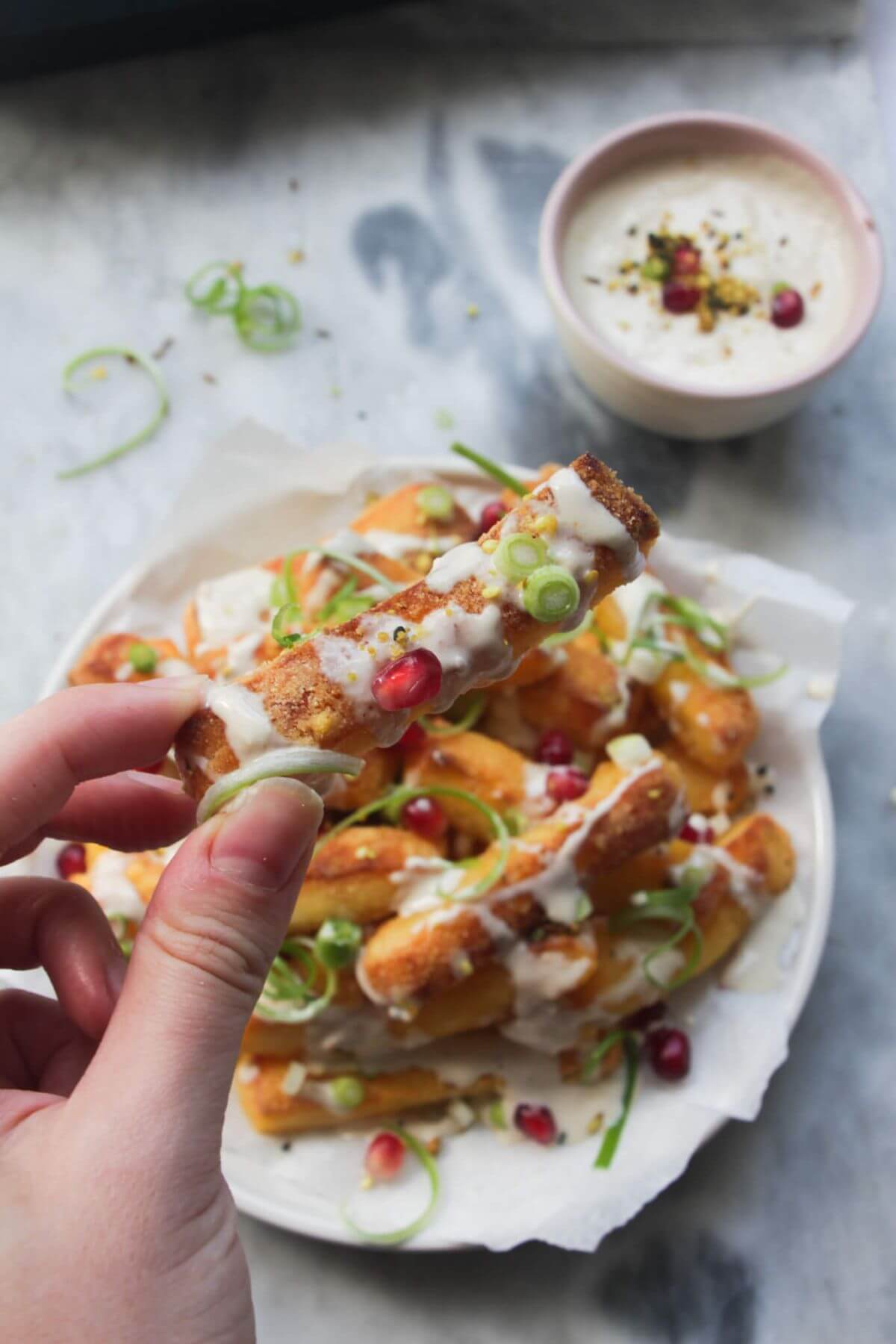 Hand holding a halloumi fry with tahini sauce, pomegratate seeds and spring onions on it.