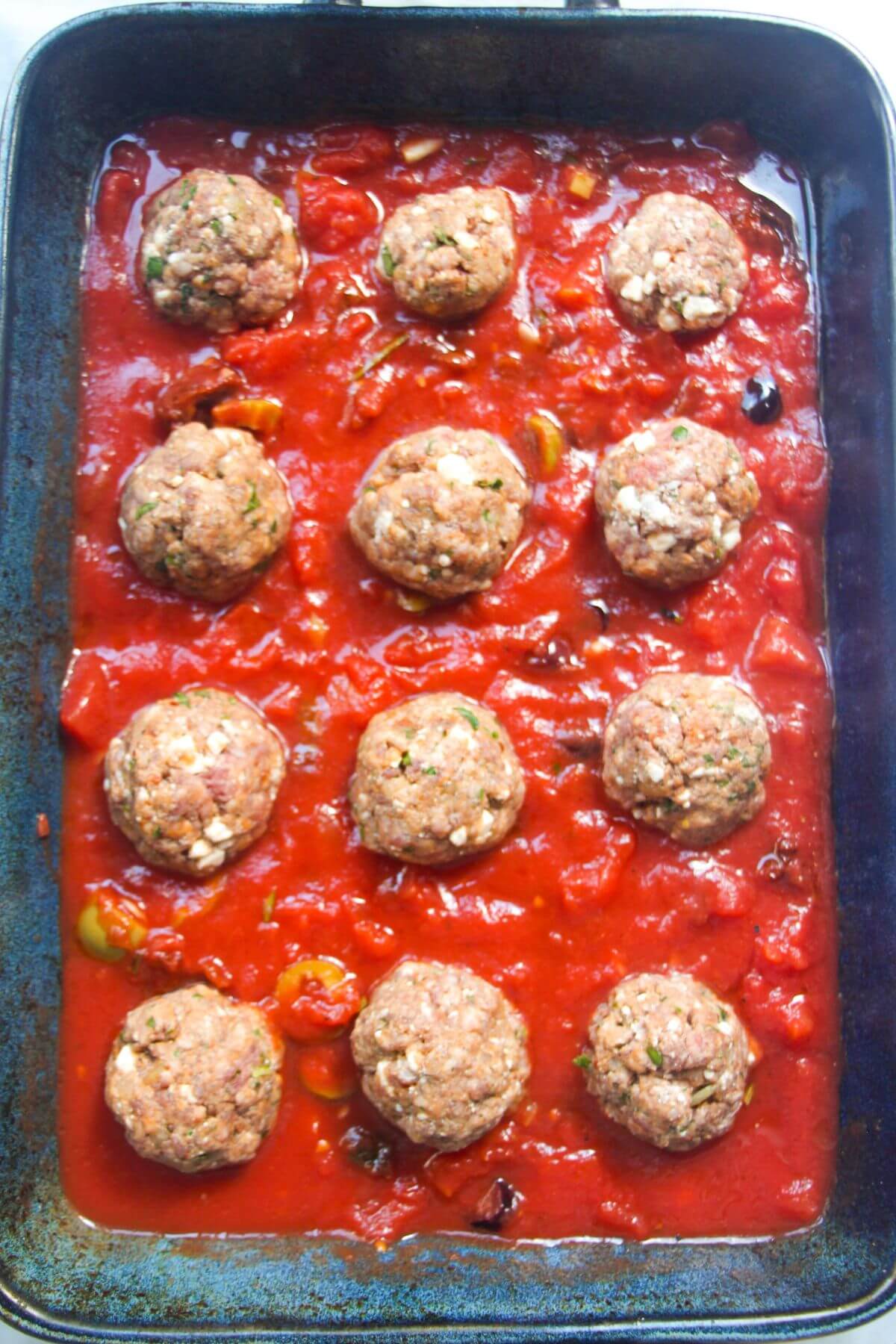 12 uncooked meatballs placed in tomato sauce in a large blue oven dish.