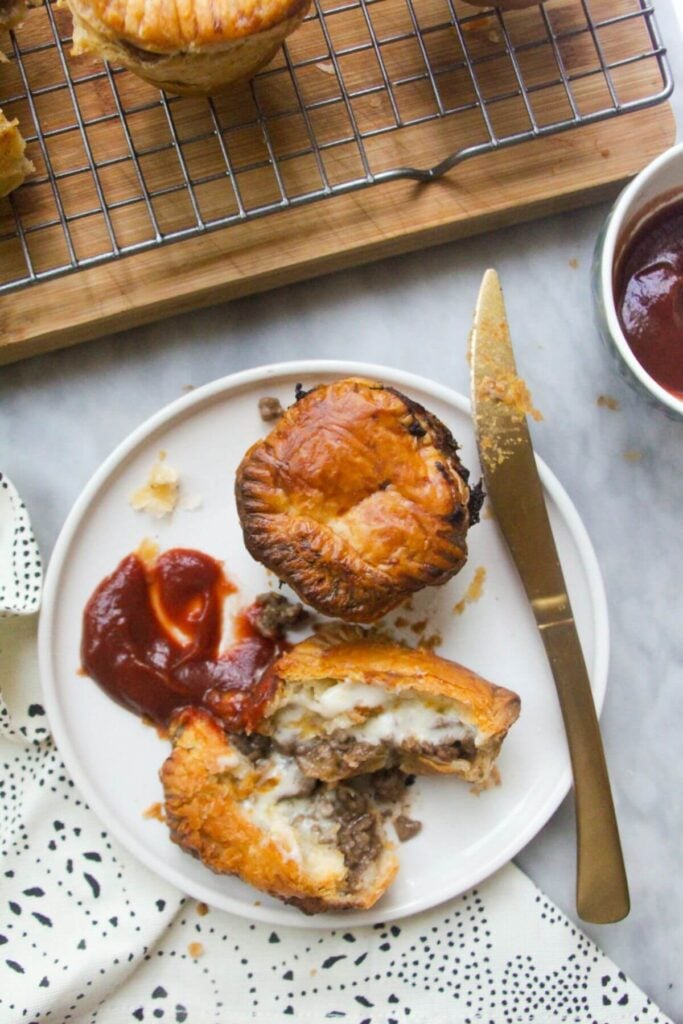 One cut open mince and cheese pie on a plate with beef and cheese filling oozing out, with tomato sauce on the side and another small mince and cheese pie on the plate behind it.