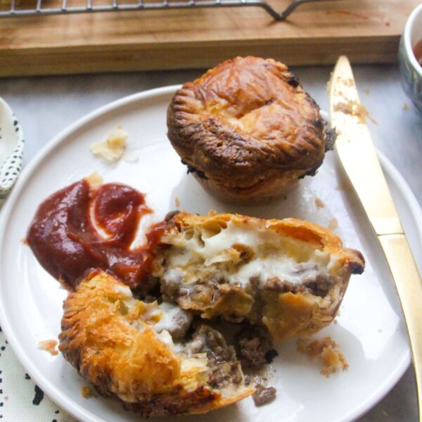 One cut open mince and cheese pie on a plate with beef and cheese filling oozing out, with tomato sauce on the side and another small mince and cheese pie on the plate behind it.