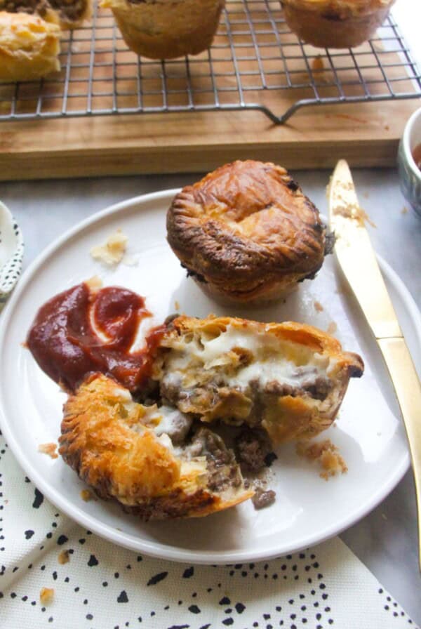 One cut open mince and cheese pie on a plate with beef and cheese filling oozing out, with tomato sauce on the side and another small mince and cheese pie on the plate behind it.