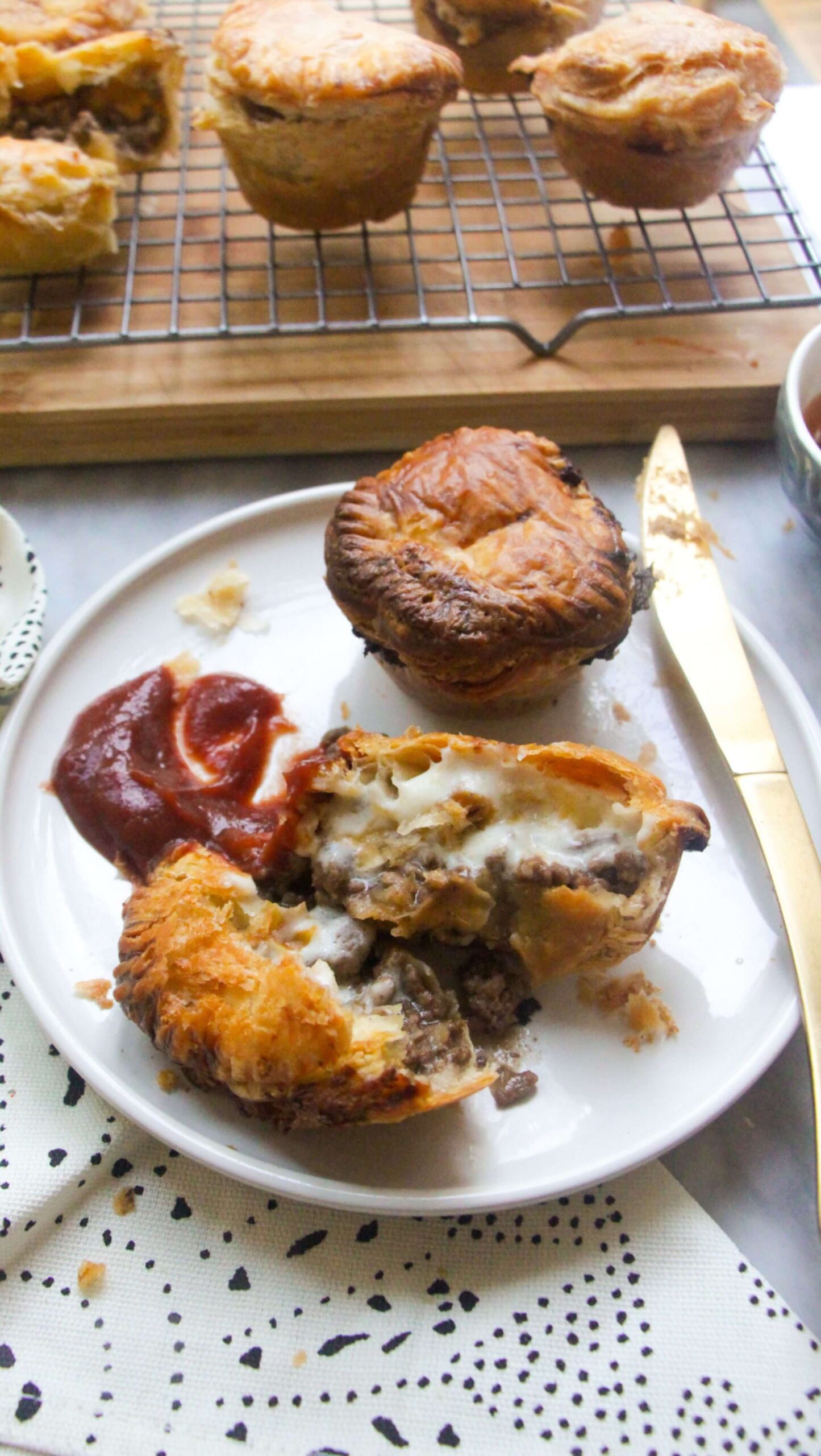 One cut open mince and cheese pie on a plate with beef and cheese filling oozing out, with tomato sauce on the side and another small mince and cheese pie on the plate behind it.