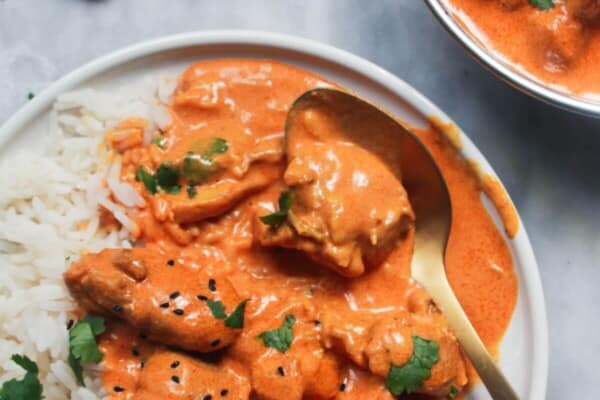 White plate with rice and butter chicken with bowl of butter chicken in the background and garlic naan.