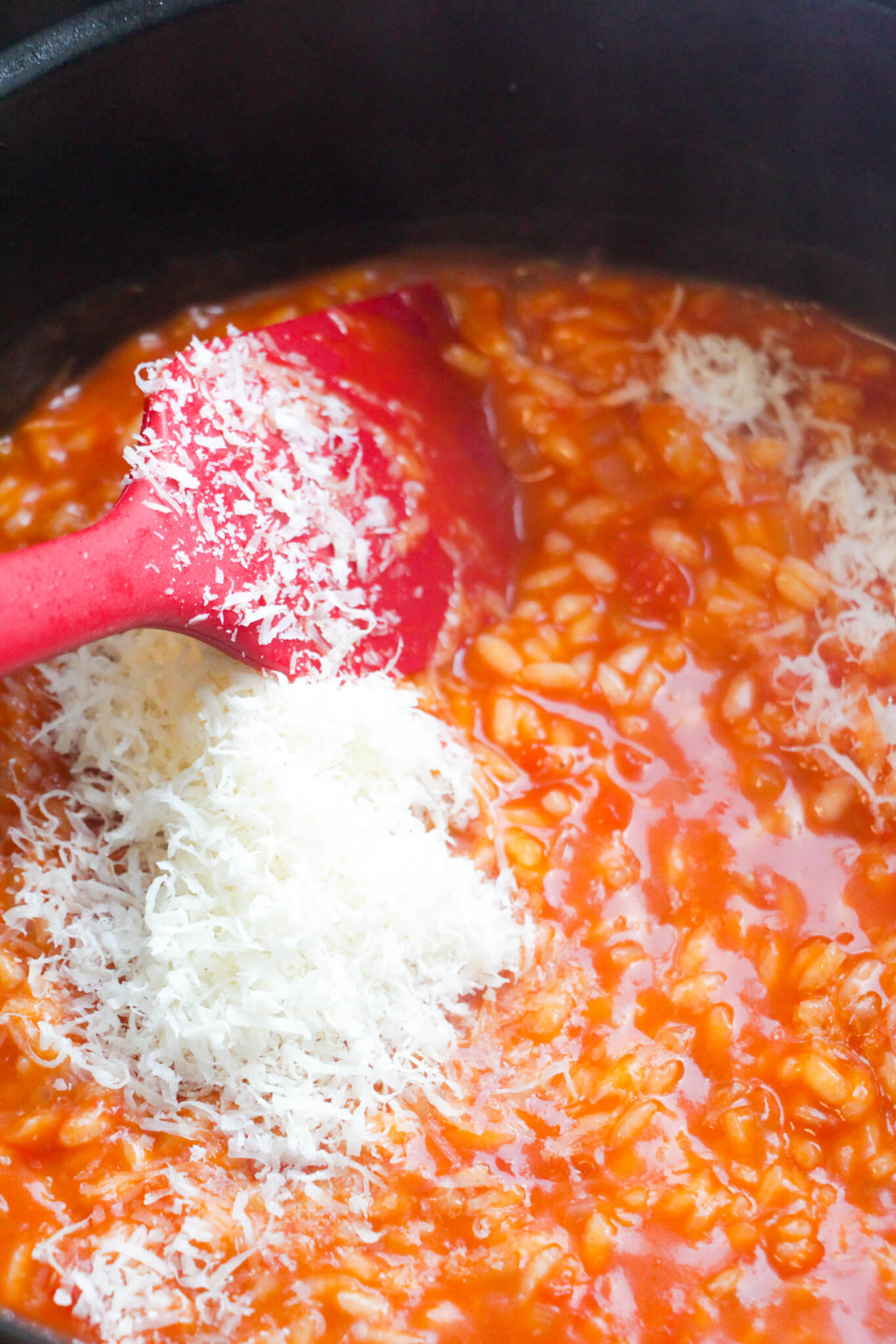 Red spatula stirring through mascarpone and parmesan in creamy tomato risotto in a black pot.