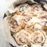 8 baked and iced cinnamon rolls in a baking paper lined cake tin with pumpkin spice in a small bowl in the background.