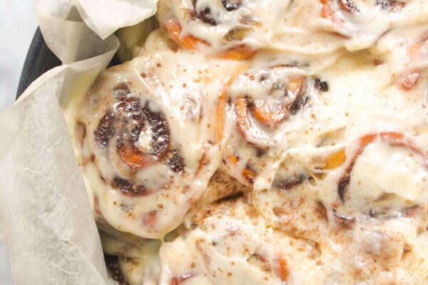 8 baked and iced cinnamon rolls in a baking paper lined cake tin with pumpkin spice in a small bowl in the background.