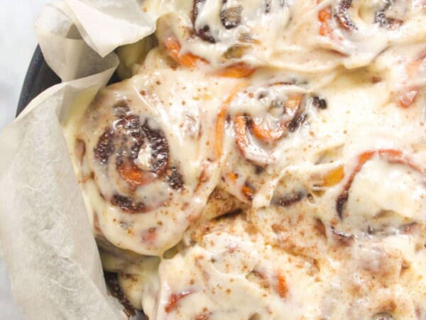 8 baked and iced cinnamon rolls in a baking paper lined cake tin with pumpkin spice in a small bowl in the background.