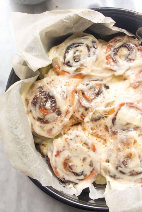 8 baked and iced cinnamon rolls in a baking paper lined cake tin with pumpkin spice in a small bowl in the background.