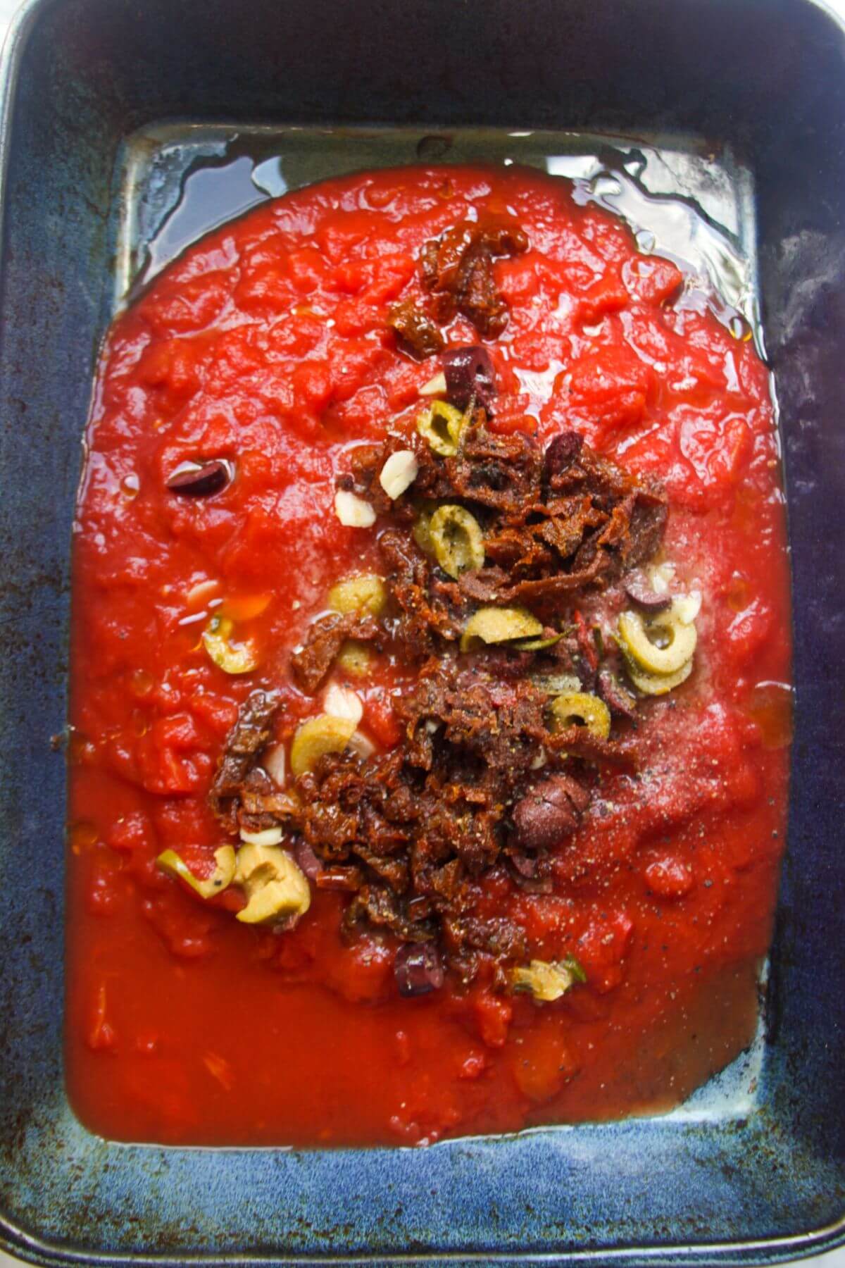Crushed tomatoes, olives and sundried tomatoes in the middle of a large blue oven dish.