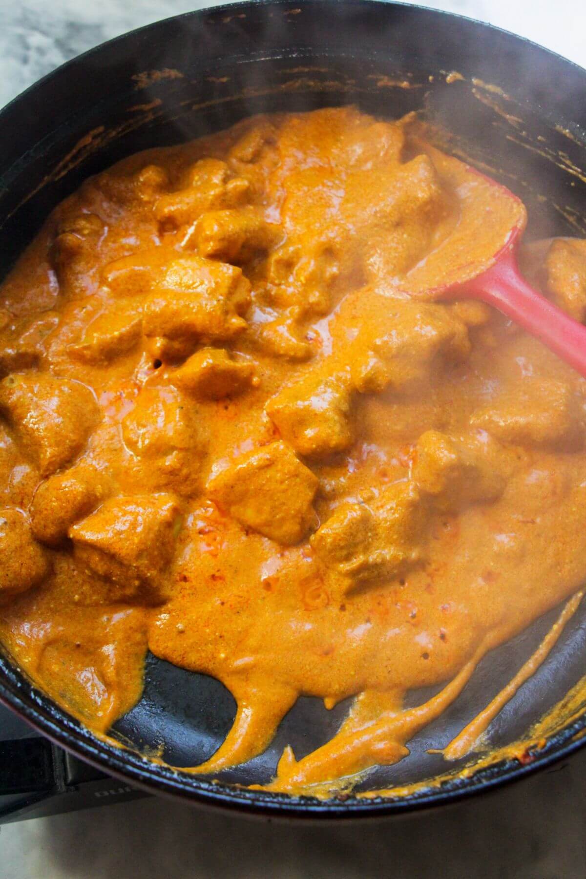 Bright orange butter chicken curry simmering in a large black pan with a red spatula.