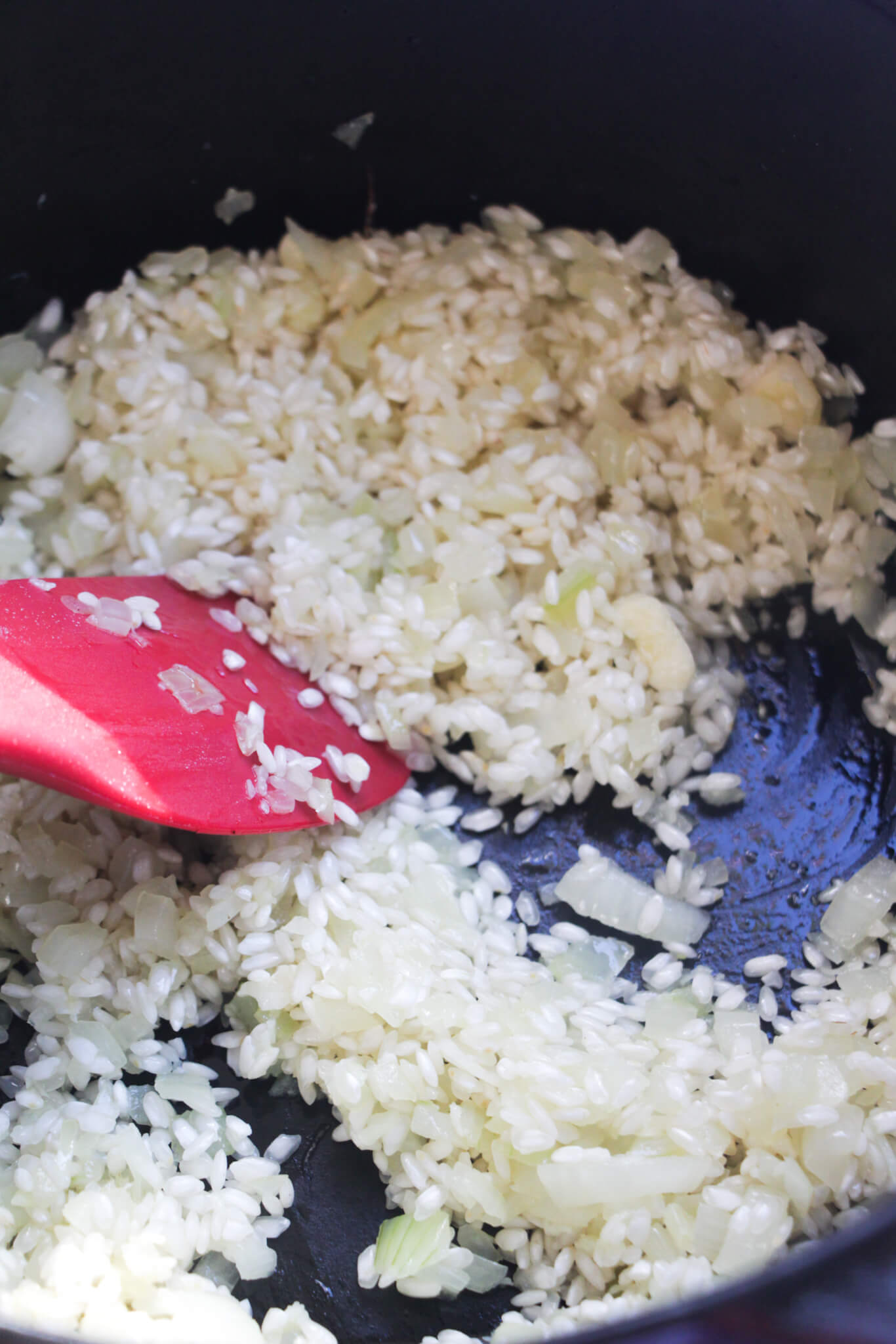 Red spatula stirring rice through onions and garlic in a large black pot.