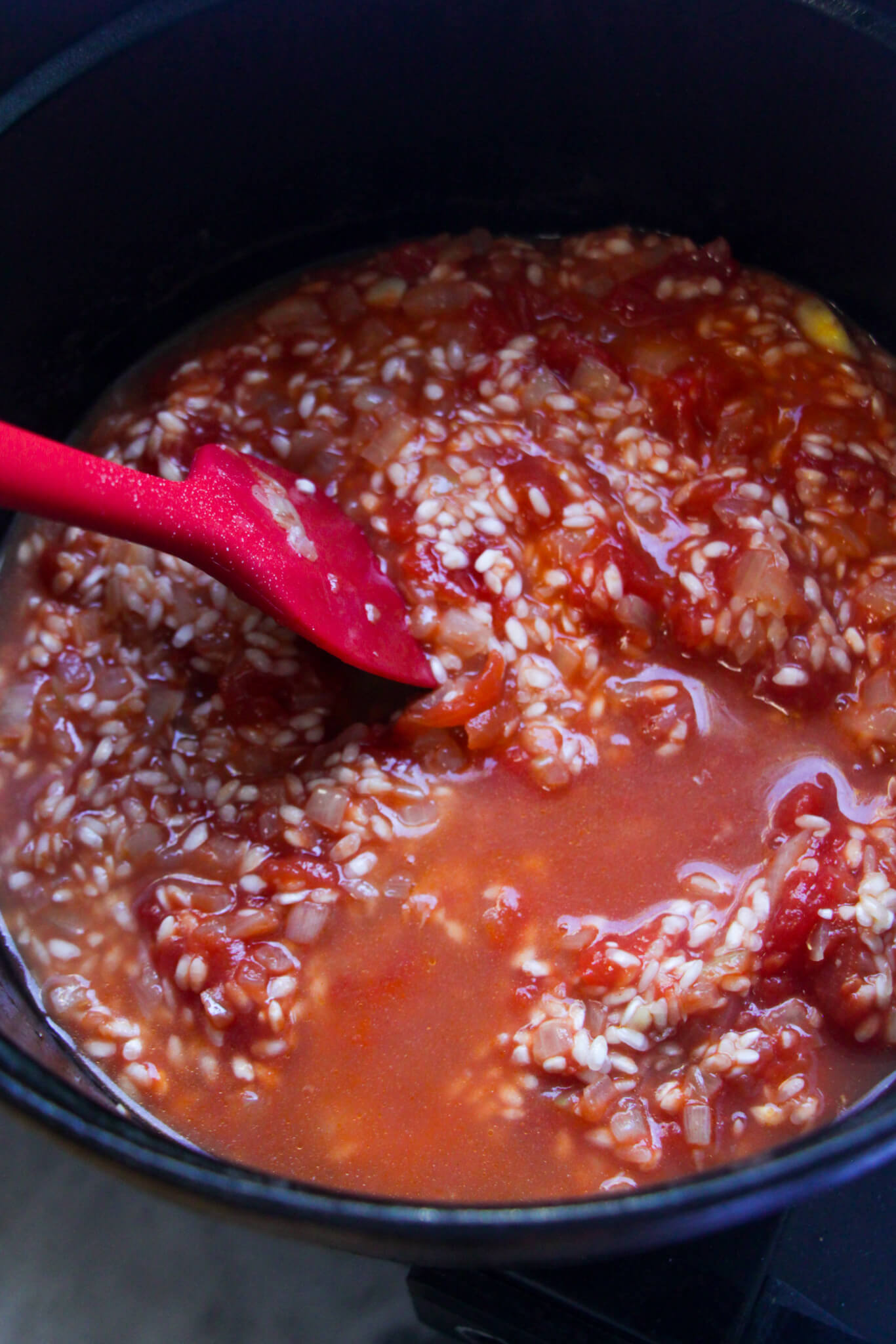Red spatula stirring stock through tomato risotto in a black pot.