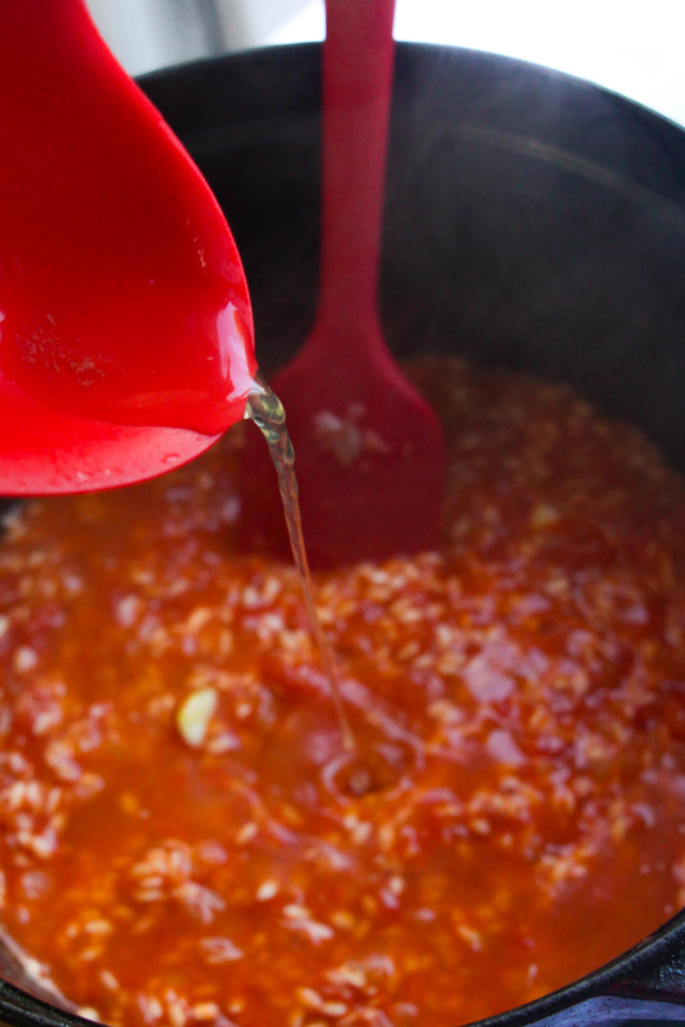 Red ladle pouring stock into tomato risotto with red spatula in the background.