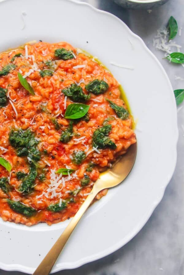 Tomato mascarpone risotto in a large white plate with dollop of green pesto on top, with a gold spoon in the risotto and bowl of mascarpone in the background.