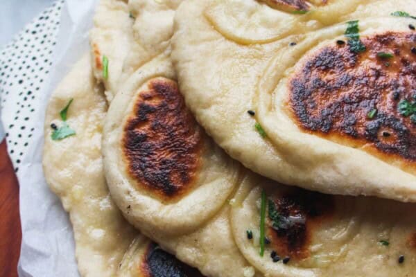 A pile of garlic naan with charred spots on baking paper on a wooden board, with a small bowl of garlic butter in the background.