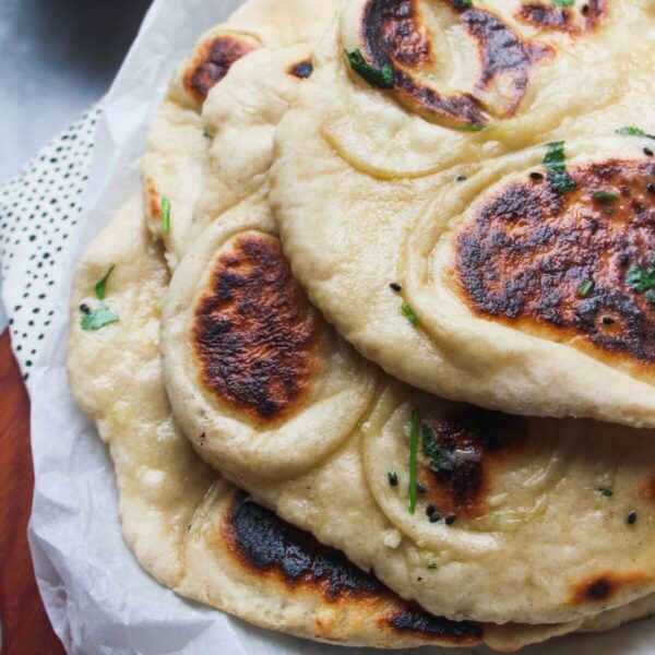 A pile of garlic naan with charred spots on baking paper on a wooden board, with a small bowl of garlic butter in the background.