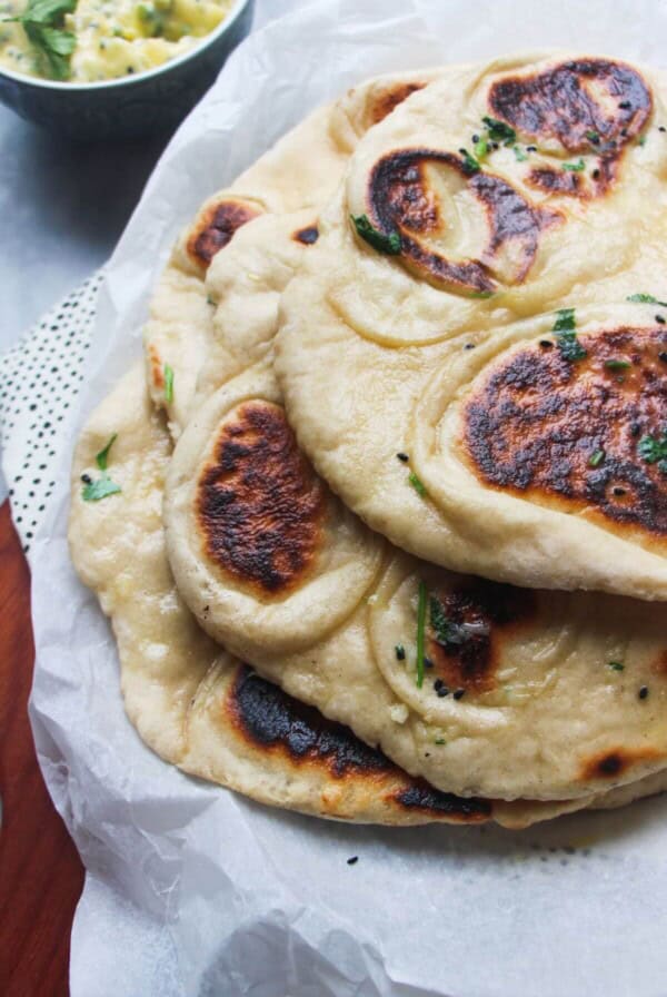 A pile of garlic naan with charred spots on baking paper on a wooden board, with a small bowl of garlic butter in the background.