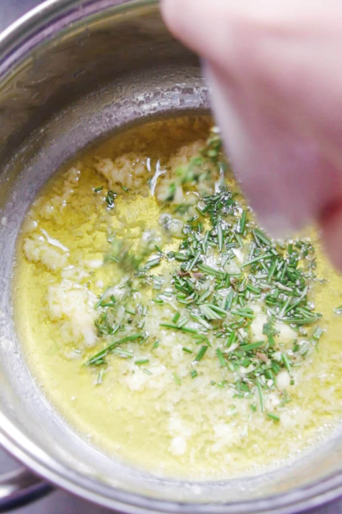 Melted butter and crushed garlic in a small silver pot, with chopped rosemary being added.