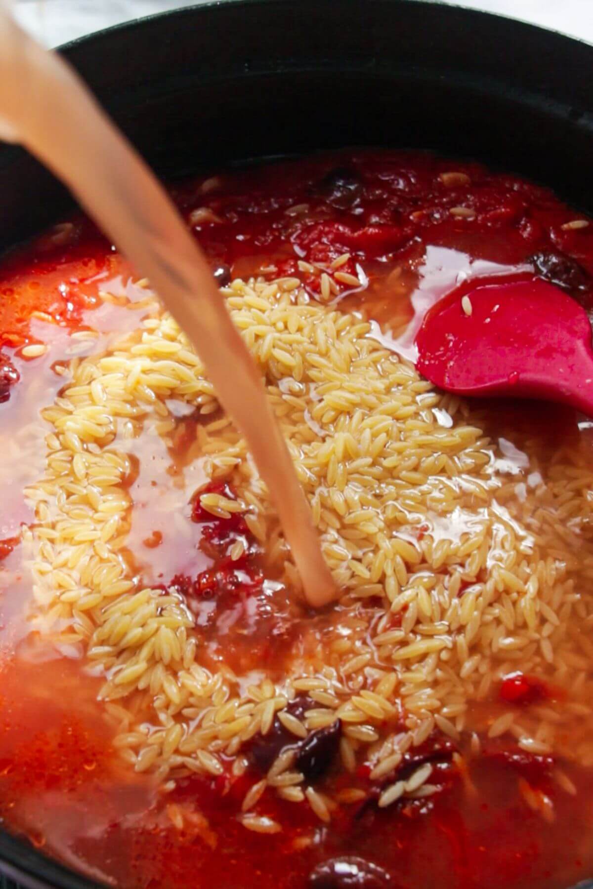 Stock being poured into orzo in a tomato sauce in a large black skillet.