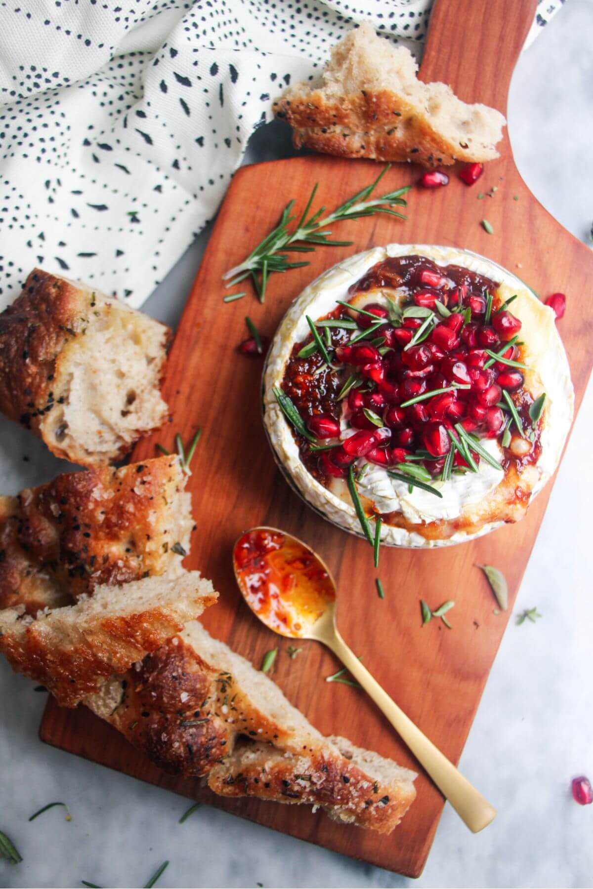 Wheel of baked camembert with pomegranate seeds, rosemary and thyme on top, with focaccia on the side.
