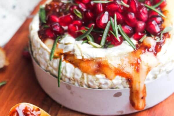 Wheel of camembert on a wooden board with oozy cheese spilling out, topped with pomegranate seeds and herbs.