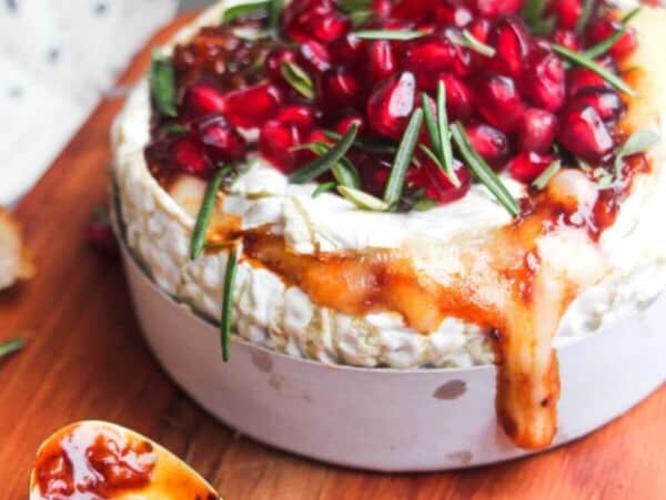 Wheel of camembert on a wooden board with oozy cheese spilling out, topped with pomegranate seeds and herbs.