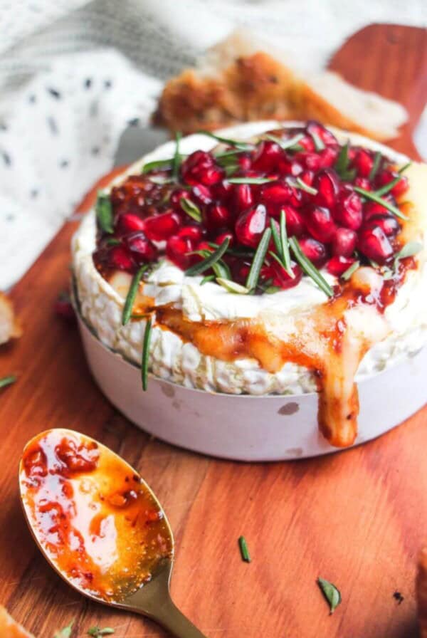 Wheel of camembert on a wooden board with oozy cheese spilling out, topped with pomegranate seeds and herbs.