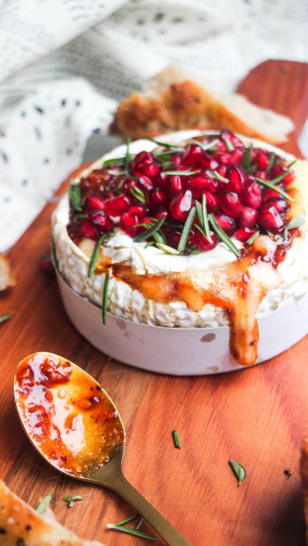 Wheel of camembert on a wooden board with oozy cheese spilling out, topped with pomegranate seeds and herbs.