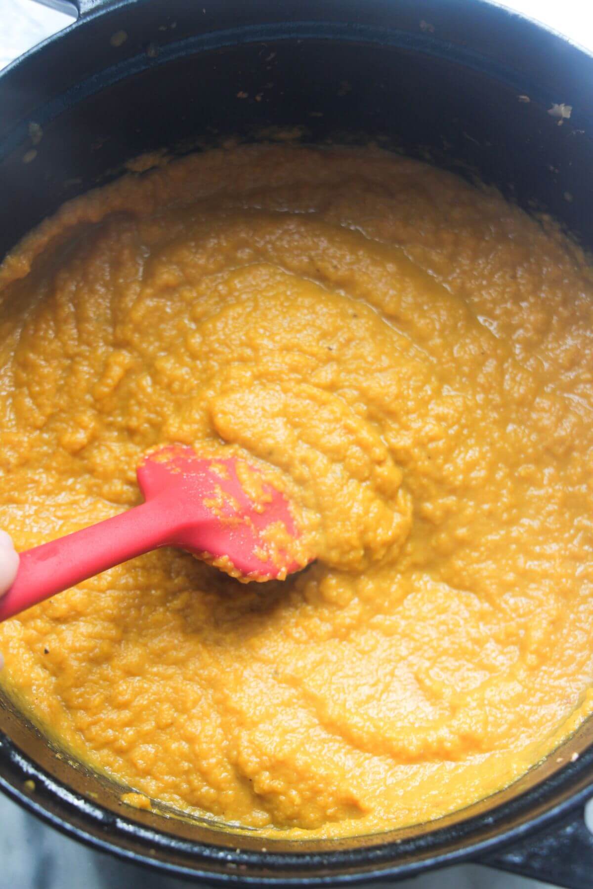 Blended carrot coriander soup in a large black pot with a red spatula stirring it.