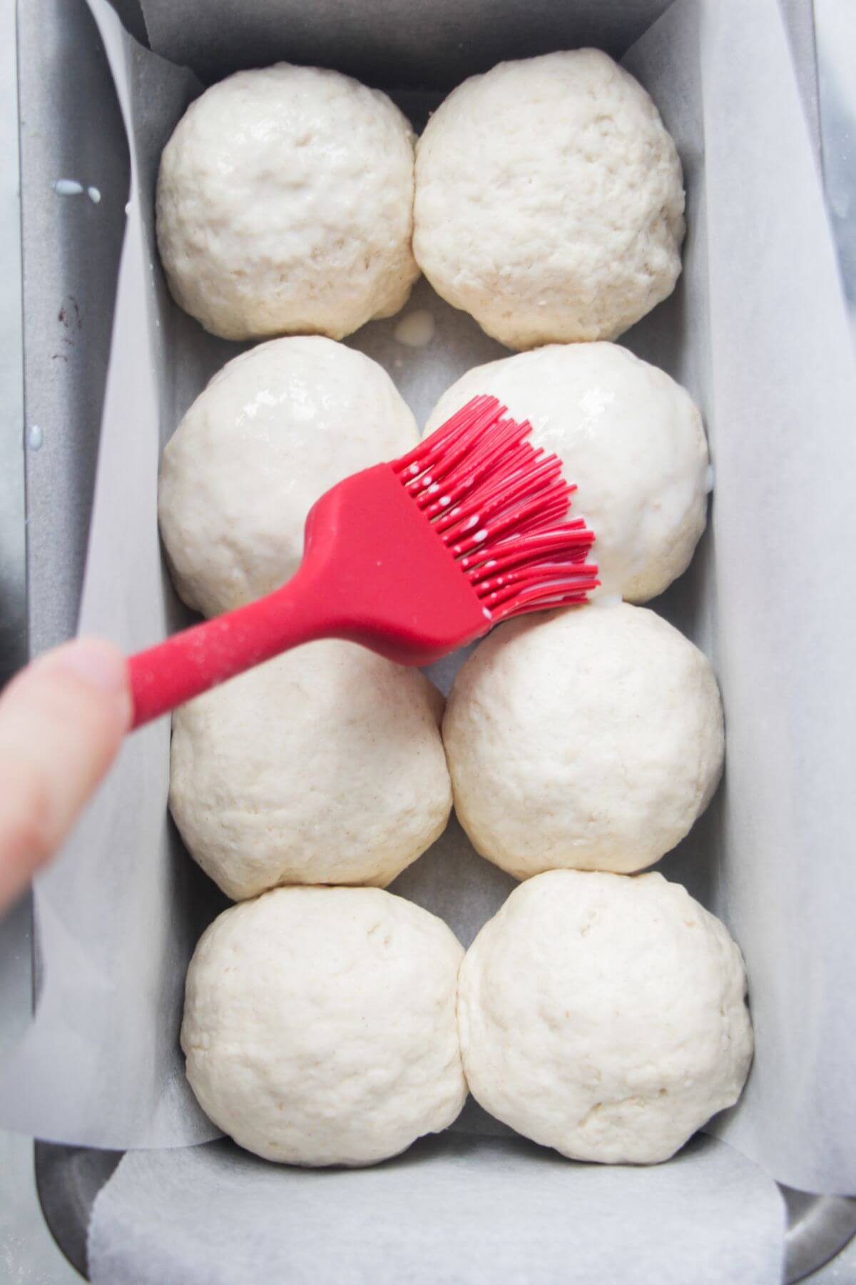 8 raw dinner rolls in a lined loaf tin being brushed with milk with a red brush.