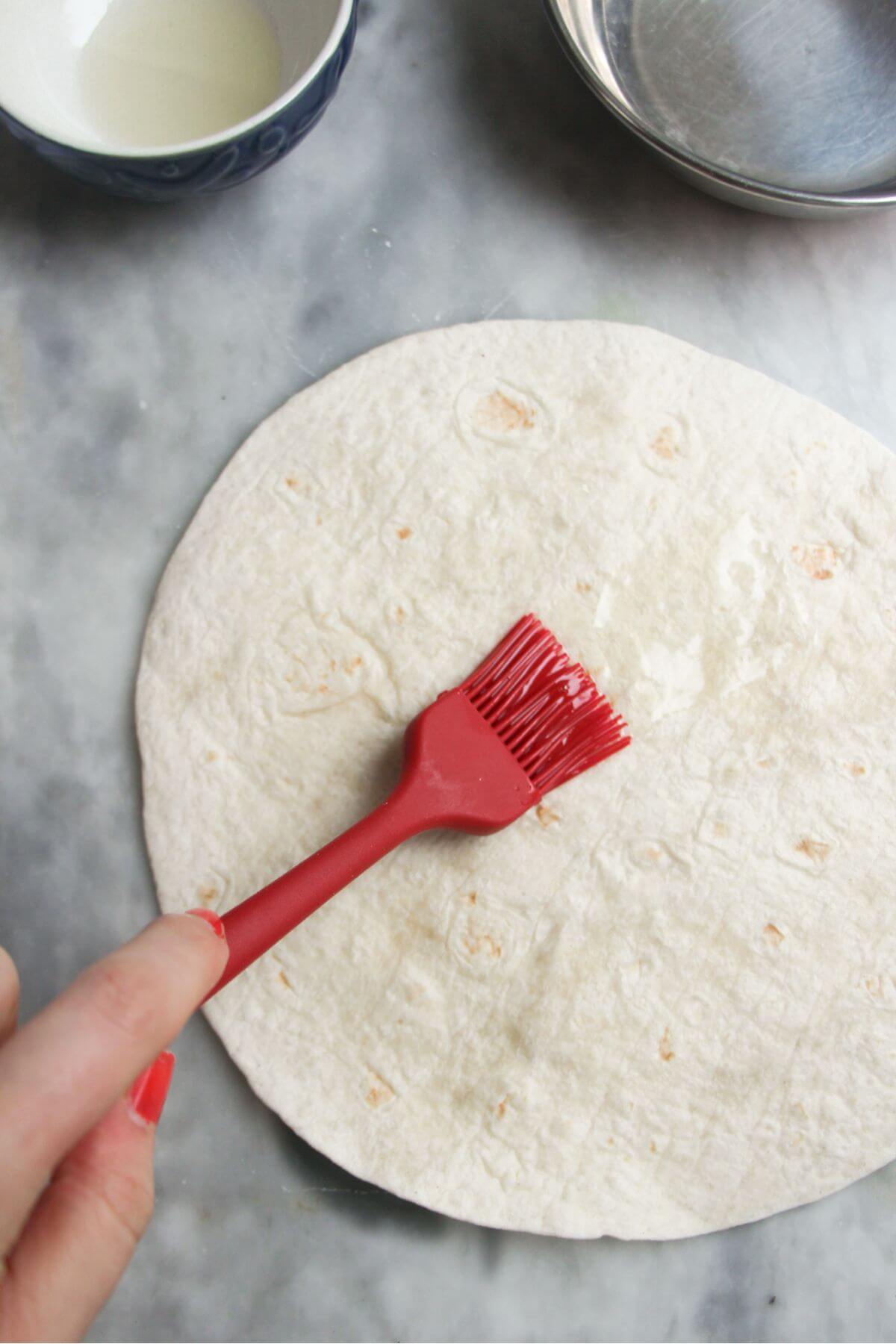 Small red brush brushing oil onto a large flour tortilla.