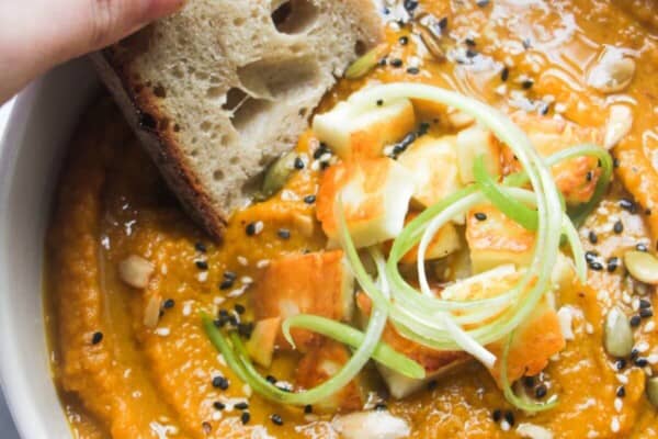 A hand dipping a slice of bread into a bowl of carrot and coriander soup with halloumi croutons.