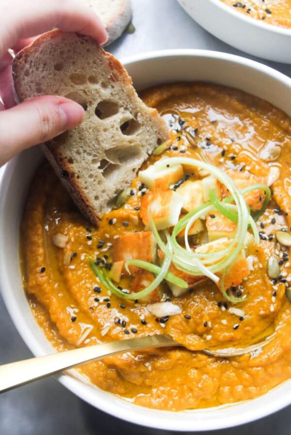 A hand dipping a slice of bread into a bowl of carrot and coriander soup with halloumi croutons.