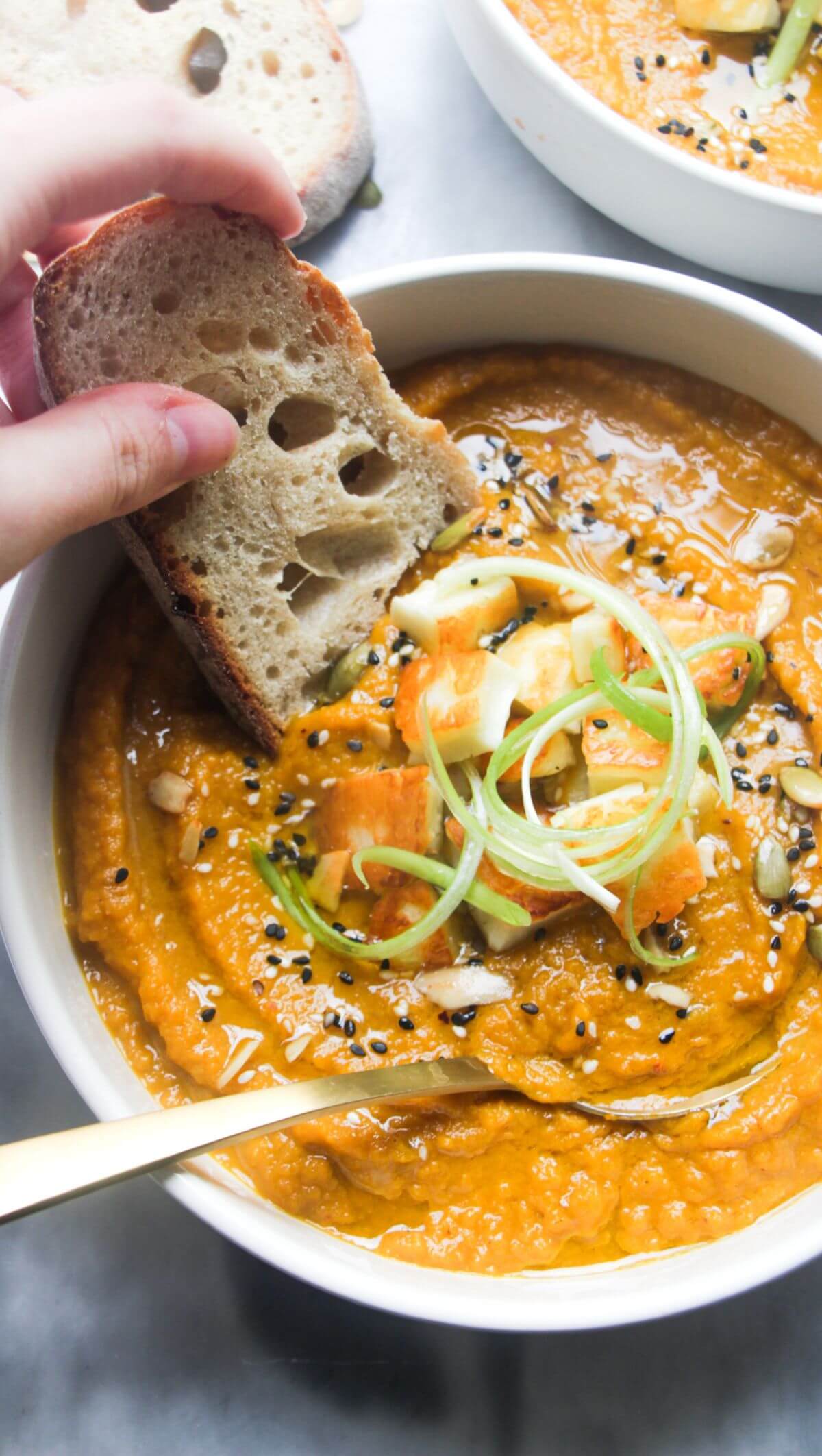 A hand dipping a slice of bread into a bowl of carrot and coriander soup with halloumi croutons.