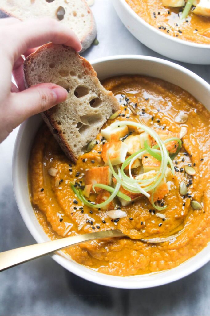 A hand dipping a slice of bread into a bowl of carrot and coriander soup with halloumi croutons.