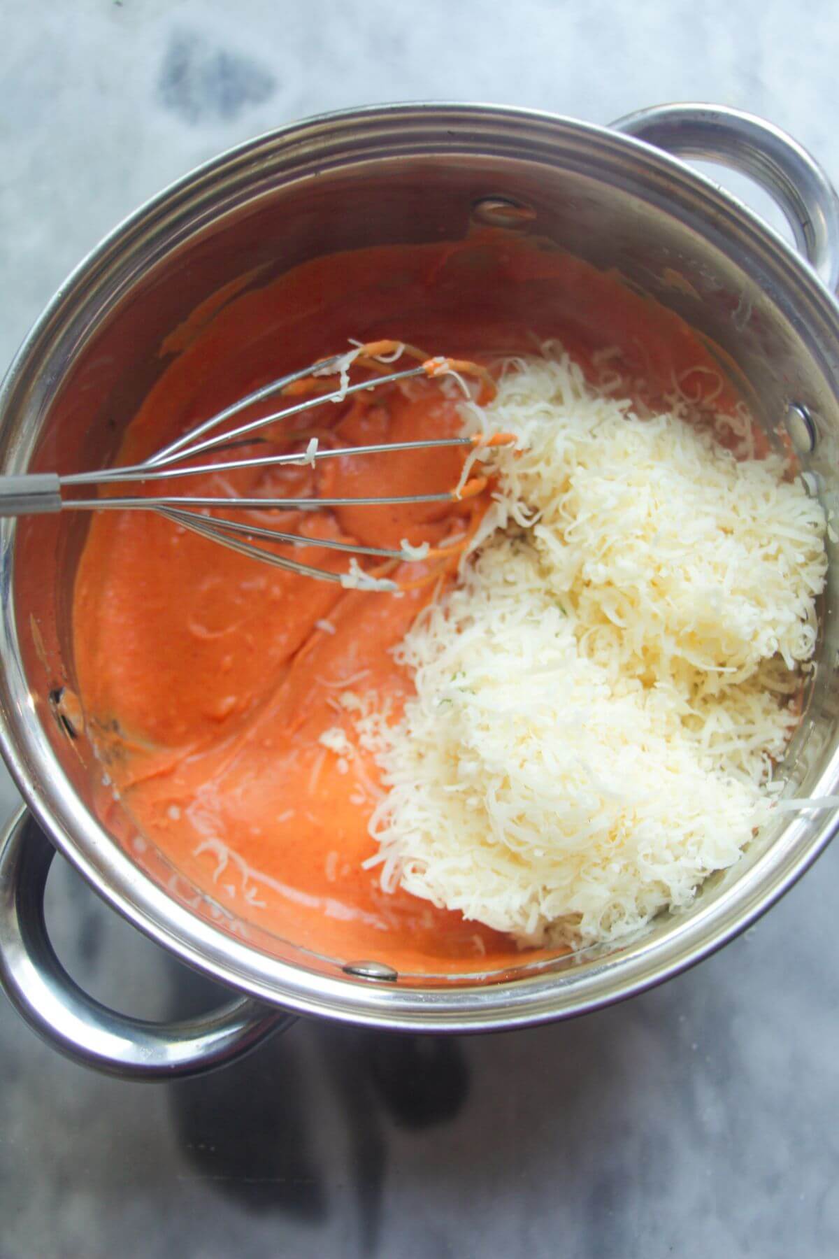 Grated cheese in a small silver pot with gochujang bechamel and a whisk inside.