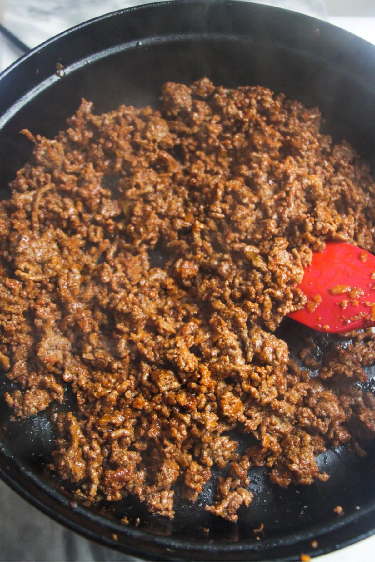 Red spatula stirring browned beef in a large black pan.