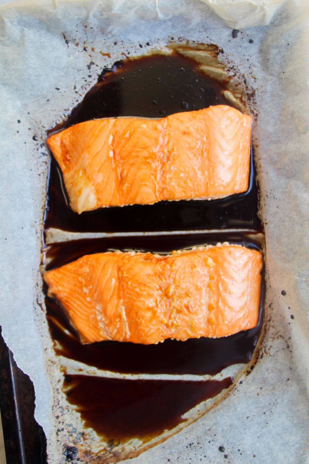 2 cooked salmon fillets sitting in soy marinade on a baking paper lined oven tray.
