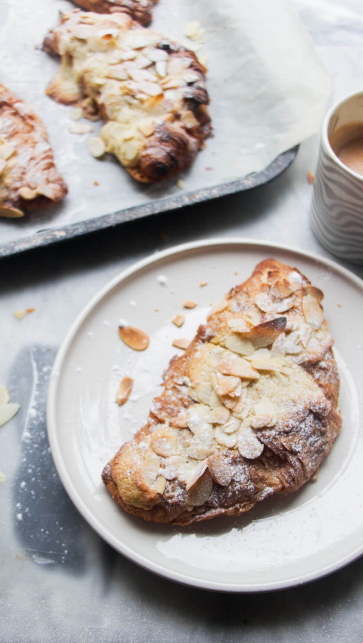 An almond croissant on a small white plate with a coffee and tray of croissants on the side.