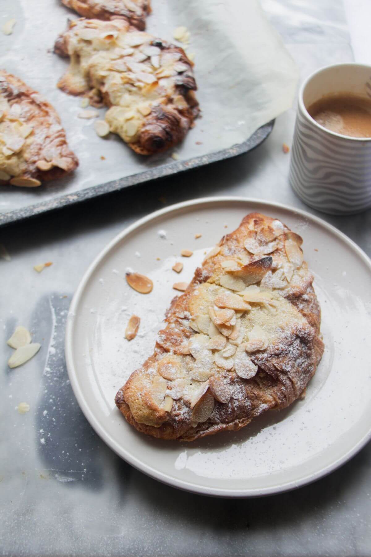 An almond croissant on a small white plate with a tray of almond croissants in the background.