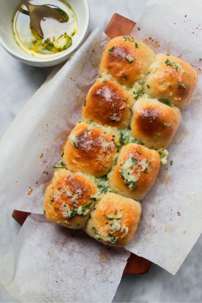 8 small rolls topped with garlic butter on a baking paper lined wooden board with bowl of garlic butter in the background.
