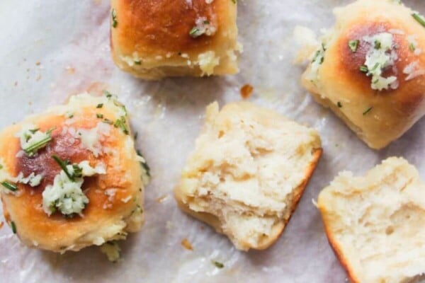 8 garlic butter topped rolled assembled on a baking paper lined wooden board with bowl of garlic butter in the background and one roll torn open.