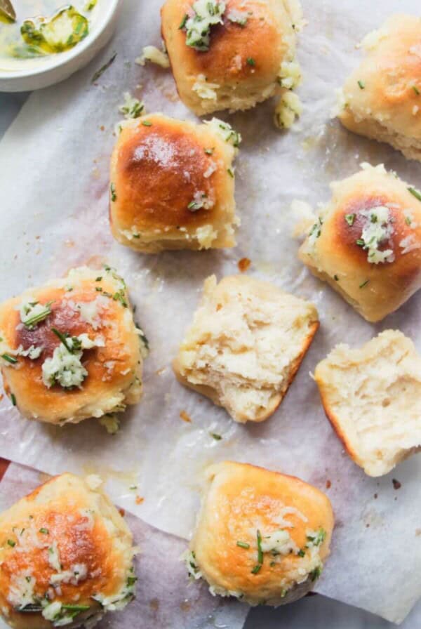8 garlic butter topped rolled assembled on a baking paper lined wooden board with bowl of garlic butter in the background and one roll torn open.