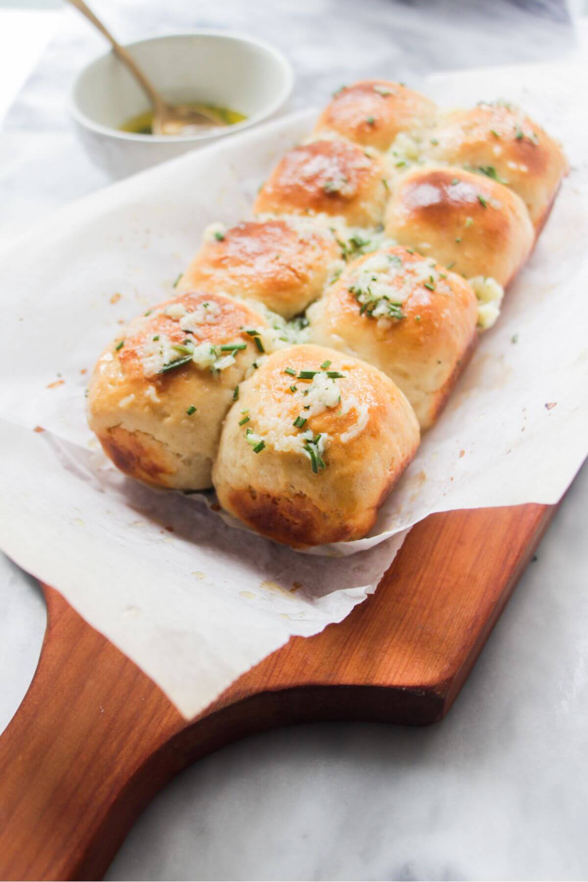 8 small rolls topped with garlic butter on a baking paper lined wooden board with bowl of garlic butter in the background.